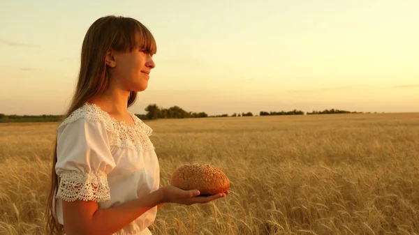 Fille tient du pain dans ses paumes et traverse le champ de blé mûr. pain dans les mains d'une jeune fille sur un champ de blé dans les rayons du coucher du soleil. savoureux pain sur les paumes . — Photo