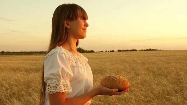 Fille tient du pain dans ses paumes et traverse le champ de blé mûr. pain dans les mains d'une jeune fille sur un champ de blé dans les rayons du coucher du soleil. savoureux pain sur les paumes . — Photo