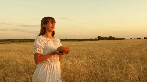 Delicioso pão nas palmas das mãos de uma jovem. menina segura pão em suas palmas e atravessa o campo de trigo maduro. pão nas mãos de uma menina sobre um campo de trigo nos raios do pôr-do-sol . — Fotografia de Stock