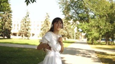 bir sırt çantası ile kız kulaklıklar Park gider ve müzik dinler ve gülümsüyor, Genç mutlu kamera objektifi elini dalgaları. Benim peşinden gel, kız şehirde seyahat ediyor. Yavaş hareket.