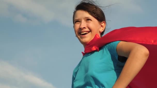 Beautiful girl superhero standing on field in red cloak, cloak fluttering in wind. Slow motion. close-up. girl dreams of becoming superhero. young girl standing in a red cloak expression of dreams. — Stock Video