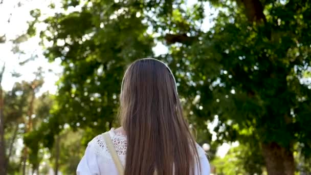 Una chica con una mochila camina por el parque con auriculares y escucha música y sonrisas agitando su mano y saludando consigo misma. chica en un vestido blanco con el pelo largo viaja por la ciudad. Movimiento lento . — Vídeos de Stock