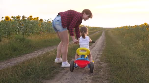 Moeder leert dochter om een fiets te rijden op een landweg in een veld van zonnebloemen. een klein kind leert fietsen. Moeder speelt met haar kleine dochter. Het concept van Happy Childhood. — Stockvideo