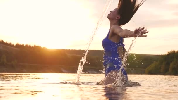 Menina brincalhão espirra seu cabelo longo em uma noite de verão refrescante perto da ilha tropical com uma água refrescante do rio. menina vira o cabelo para trás. belo spray de água em raios de pôr do sol. respingo de água — Vídeo de Stock