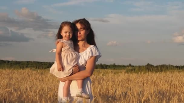 お母さんと小さな娘は熟した小麦の畑を歩きます。幸せな母親は野原で赤ちゃんと一緒に歩きます。幸せな家族旅行。お母さんの腕の中の赤ちゃんが出て行く。幸せな家族の概念. — ストック動画
