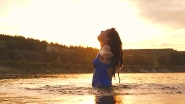 Uzun saçlı genç bir kız gün batımında yıkanır ve su sıçratıyor. Mutlu ve gülen kadın nehirde soğur. Yaz tatilleri. Sahilde hafta sonu. Özgürlük ve bağımsızlık. Yaşam tarzı. Yavaş çekim.