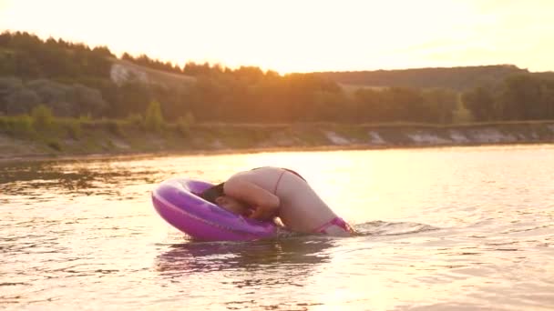 Hermosa y feliz chica en un traje de baño se sumerge en un círculo inflable en el agua y se ríe. Vacaciones de verano en la playa. Juegos en el agua. Fin de semana fuera de la ciudad — Vídeo de stock