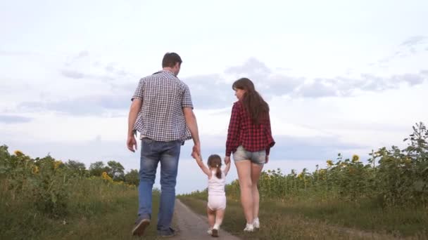 Family with small child walks along road and laughs next to field of sunflowers. Child is riding in arms of his father and mother. Mom, dad and daughter are resting together outside city in nature. — Stock Video