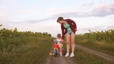 Annem kızıbir kır yolda ayçiçeği bir alanda bisiklete binmek öğretir. küçük bir çocuk bisiklete binmeyi öğrenir. Anne küçük kızıyla oynuyor. Mutlu çocukluk kavramı.