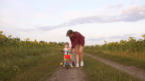 Moeder leert dochter om een fiets te rijden op een landweg in een veld van zonnebloemen. een klein kind leert fietsen. Moeder speelt met haar kleine dochter. Het concept van Happy Childhood. — Stockfoto