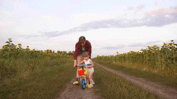 Mamma lär dotter att rida en cykel på en landsväg i ett fält av solrosor. ett litet barn lär sig att cykla. Mamma leker med sin lilla dotter. Begreppet lycklig barndom. — Stockfoto