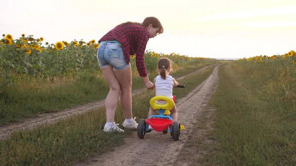Mom teaches daughter to ride a bike on a country road in a field of sunflowers. a small child learns to ride a bike. Mother plays with her little daughter. The concept of happy childhood.
