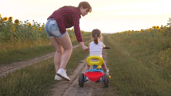 Moeder leert dochter om een fiets te rijden op een landweg in een veld van zonnebloemen. een klein kind leert fietsen. Moeder speelt met haar kleine dochter. Het concept van Happy Childhood. — Stockfoto