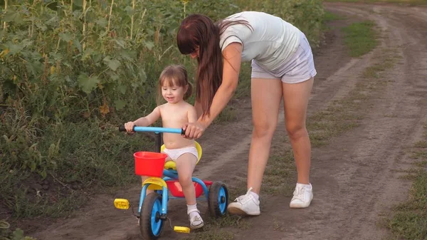 ママは娘に自転車に乗るように教える。妹は小さな子供と遊ぶ。幸せな子供時代の概念。小さな子供は自転車に乗ることを学ぶ. — ストック写真