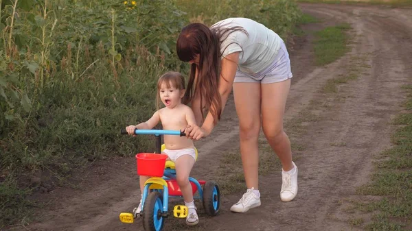 Mamá enseña a su hija a andar en bicicleta. La hermana juega con un niño pequeño. El concepto de la infancia feliz. un niño pequeño aprende a andar en bicicleta . — Foto de Stock