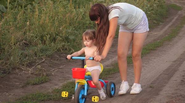 ママは娘に自転車に乗るように教える。妹は小さな子供と遊ぶ。幸せな子供時代の概念。小さな子供は自転車に乗ることを学ぶ. — ストック写真