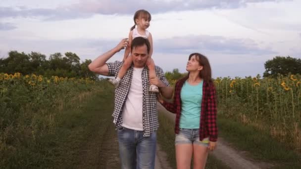 Family with small baby is walking along road and laughing next to field of sunflowers. Child is riding on his father shoulders. Mom, dad and daughter are resting together outside city in nature. — Stock Video