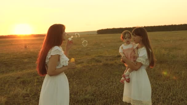 Döttrar gläds och ler, bubblor flyger i parken vid solnedgången. Slow motion. Lycklig mamma leker med barn blåser tvål bubblor. konceptet med en lycklig familj. barn, Syster och mor spelar på Sunrise. — Stockvideo