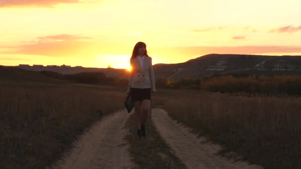 Sexy mulher de negócios andando ao longo de uma estrada rural com uma pasta na mão, o sol brilha ao pôr do sol. Menina de negócios sexy trabalhando no campo. menina bonita viaja o campo . — Vídeo de Stock