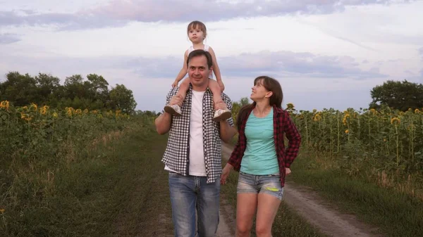 Familj med små barn går längs vägen och skrattar bredvid fältet av solrosor. Barnet rider på sin fars axlar. Mamma, Pappa och dotter vilar tillsammans utanför staden i naturen. — Stockfoto