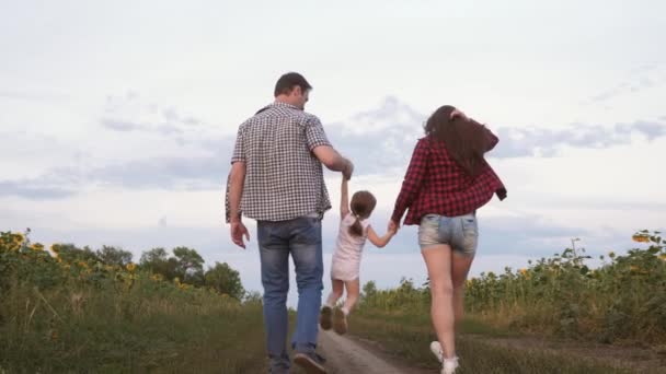 Little daughter jumping holding hands mom and dad. Family with small child walks along road and laughs next to field of sunflowers. Mom, dad and daughter are resting together outside city in nature — Stock Video