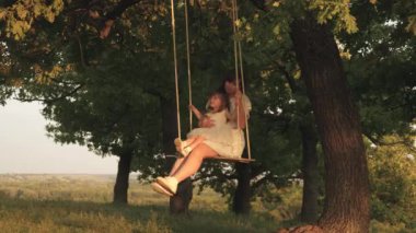 anne ve bebek ormanda bir meşe dalı üzerinde bir ip salıncak binmek. Anne güneşte bir ağacın altında salıncakta kızını sallar. Kız güler, sevinir. Parkta, doğada aile eğlencesi. sıcak yaz günü.