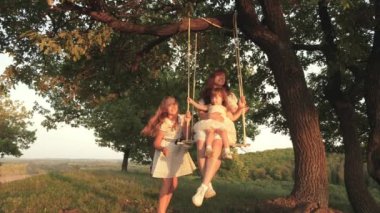 Anne, güneşte ağacın altında salıncakta kızlarını sallıyor, çocuklarla oynuyor. anne ve bebek ormanda bir meşe dalı üzerinde halat salıncak binmek. Kız güler, sevinir. Parkta, doğada aile eğlencesi