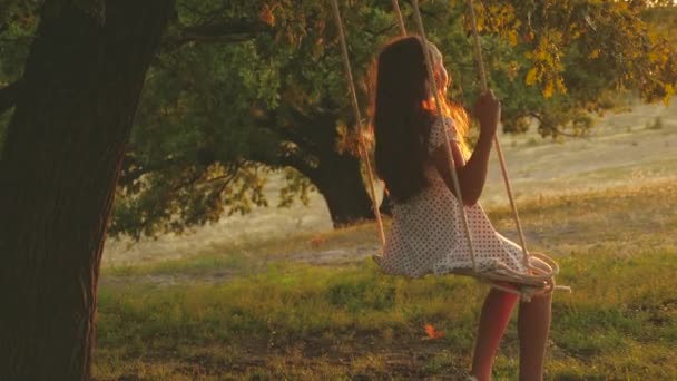 在公园日落时分，孩子骑着绳子在橡树树枝上摆动。特写。女孩笑了，高兴。小女孩在阳光下在树下秋千上摇摆，和孩子们玩耍。大自然中的家庭乐趣。慢动作 — 图库视频影像