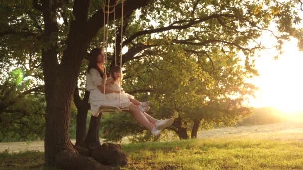 Mamma scuote sua figlia sull'altalena sotto un albero al sole. madre e bambino cavalcano su un'altalena di corda su un ramo di quercia nella foresta. La ragazza ride, gioisce. Divertimento in famiglia nel parco, nella natura. calda giornata estiva . — Video Stock