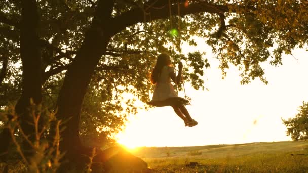 Kind rijdt een touwschommel op een eiken tak in bos. meisje lacht, verheugt. jong meisje swingende op een schommel onder een boom in de zon, spelen met kinderen. Close-up. Familie plezier in Park, in de natuur. — Stockvideo