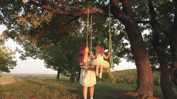 Mamá sacude a su hija en columpio bajo un árbol bajo el sol. niño ríe y se regocija. madre y el bebé montar en un columpio cuerda en una rama de roble en el bosque. Diversión familiar en el parque, en la naturaleza. cálido día de verano . — Vídeo de stock