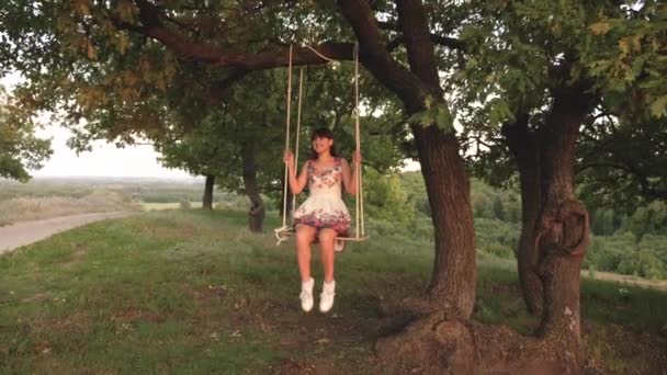 Jong meisje swingende op een touwschommel op een eiken tak. Mooi meisje in een witte jurk in het Park. tiener meisje geniet van een vlucht op een schommel op een zomeravond in het bos — Stockvideo