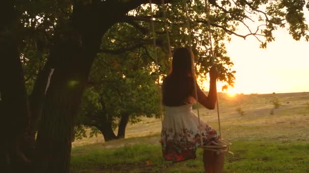 Teen girl gode di un volo su un'altalena in una serata estiva nella foresta. giovane ragazza che oscilla su un'altalena di corda su un ramo di quercia. Bella ragazza in un vestito bianco nel parco . — Video Stock