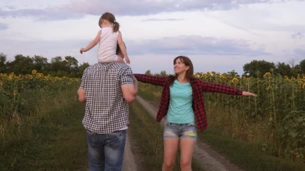 Rodzina z małym dzieckiem jest chodzenie wzdłuż drogi i śmiech obok pola słoneczniki. Dziecko jeździ na ramionach ojca. Mama, Tata i córka odpoczywają razem poza miastem w przyrodzie. — Wideo stockowe
