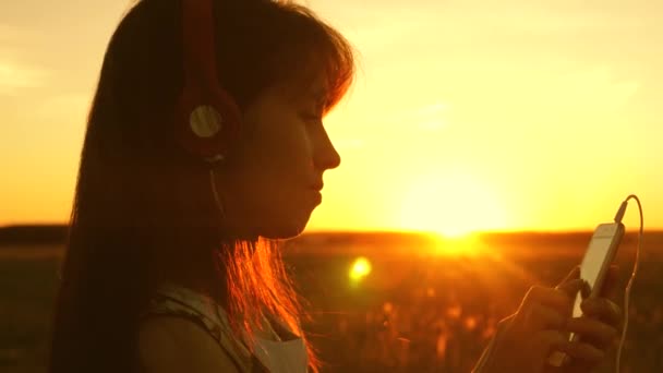 Viaggiatore di ragazza che ascolta musica su smartphone in raggi di un bel tramonto di primavera. ragazza con le cuffie e con tablet seleziona canzoni online, in parco in estate in raggi di sole . — Video Stock