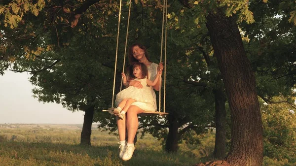 Madre y el bebé montar en un columpio cuerda en una rama de roble en el bosque. Mamá sacude a su hija en columpio bajo un árbol bajo el sol. La chica se ríe, se alegra. Diversión familiar en el parque, en la naturaleza. cálido día de verano . —  Fotos de Stock