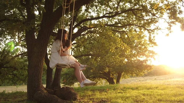 Mamma skakar sin dotter på swing under ett träd i solen. mor och baby rida på ett rep Swing på en ek gren i skogen. Flickan skrattar, jublar. Familj kul i parken, i naturen. varma sommardagen. — Stockfoto