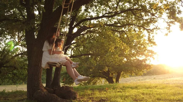 Mutter schüttelt ihre Tochter auf Schaukel unter einem Baum in der Sonne. Mutter und Baby reiten auf einer Seilschaukel auf einem Eichenzweig im Wald. Mädchen lacht, freut sich. Familienspaß im Park, in der Natur. Warmer Sommertag. — Stockfoto