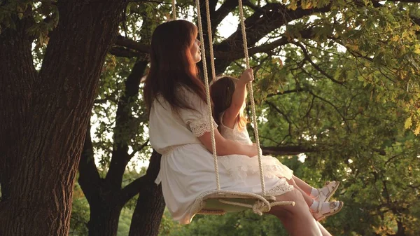 Mamá sacude a su hija en columpio bajo un árbol bajo el sol. Primer plano. madre y el bebé montar en un columpio cuerda en una rama de roble en el bosque. La chica se ríe, se alegra. Diversión familiar en el parque, en la naturaleza. cálido día de verano . —  Fotos de Stock