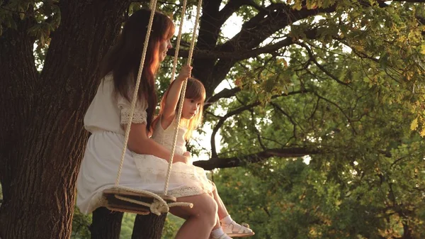 Mamma skakar sin dotter på swing under ett träd i solen. Närbild. mor och baby rida på ett rep Swing på en ek gren i skogen. Flickan skrattar, jublar. Familj kul i parken, i naturen. varma sommardagen. — Stockfoto