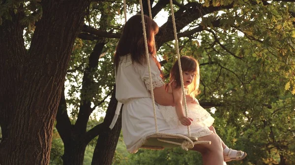 妈妈在阳光下摇摇着女儿在树下。特写。母亲和婴儿骑在森林的橡树树枝上的绳索摆动。女孩笑了，高兴。家庭乐趣在公园，在自然。温暖的夏日. — 图库照片