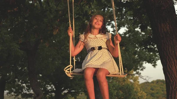 Ung flicka svänger på en gunga under ett träd i solen, leker med barn. Närbild. barn rider ett rep Swing på en ek gren i skogen. flickan skrattar, jublar. Familj kul i parken, i naturen. — Stockfoto