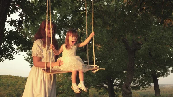 Mamma skakar sin dotter på swing under Tree in Sun. Närbild. barn skrattar och jublar. mor och baby rida på ett rep Swing på en ek gren i skogen. Familj kul i parken, i naturen. varma sommardagen — Stockfoto