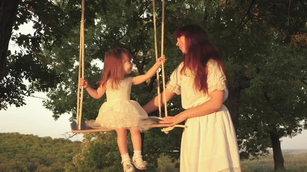 Maman secoue sa fille en se balançant sous un arbre au soleil. Gros plan. enfant rit et se réjouit. mère et bébé montent sur une balançoire à corde sur une branche de chêne dans la forêt. Amusement en famille dans le parc, dans la nature. chaude journée d'été — Photo