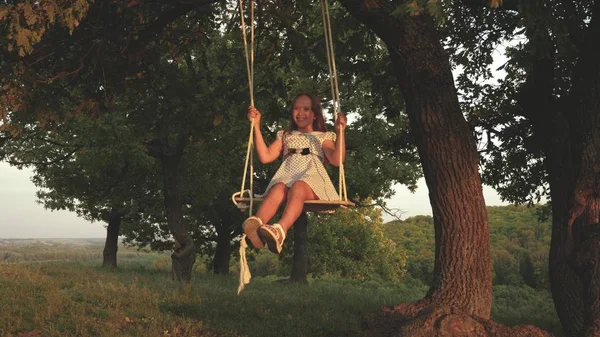 Vacker ung flicka svänger på en gunga under ett träd i solen, leker med barn. barn rider ett rep Swing på en ek gren i skogen. flickan skrattar, jublar. Familjen kul i parken, i naturen. — Stockfoto