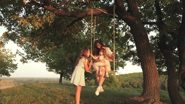 Mama schüttelt ihre Töchter auf einer Schaukel unter einem Baum in der Sonne und spielt mit Kindern. Mutter und Baby reiten auf einer Seilschaukel auf einem Eichenzweig im Wald. Mädchen lacht, freut sich. Familienspaß im Park, in der Natur — Stockfoto