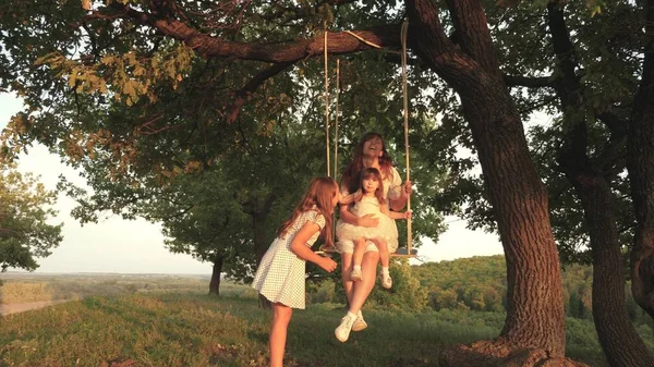 Mamãe balança suas filhas em um balanço debaixo da árvore ao sol, brincando com as crianças. mãe e bebê passeio na corda balançar em um galho de carvalho na floresta. Ri menina, alegra-se. Diversão familiar no parque, na natureza — Fotografia de Stock