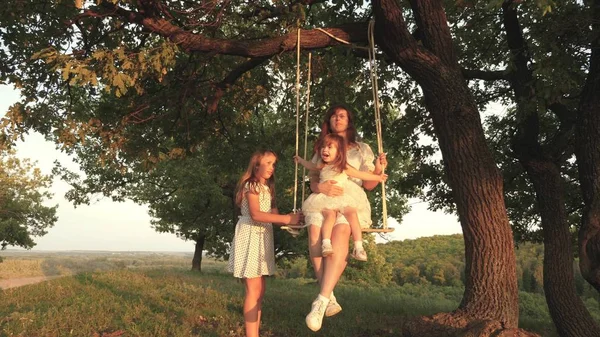 Mamma skakar sina döttrar på en gunga underträd i solen, leka med barn. mor och baby rida på rep Swing på en ek gren i skogen. flickan skrattar, jublar. Familj kul i parken, i naturen — Stockfoto