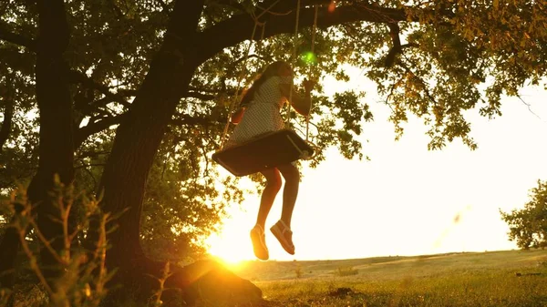 Niño monta un columpio de cuerda en una rama de roble en el bosque. chica ríe, se regocija. jovencita balanceándose en un columpio bajo un árbol en el sol, jugando con los niños. Primer plano. Diversión familiar en el parque, en la naturaleza . — Foto de Stock