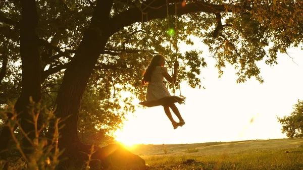 Niño monta un columpio de cuerda en una rama de roble en el bosque. chica ríe, se regocija. jovencita balanceándose en un columpio bajo un árbol en el sol, jugando con los niños. Primer plano. Diversión familiar en el parque, en la naturaleza . —  Fotos de Stock